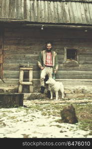 handsome young hipster man standing together with white husky dog in front of old vintage retro wooden house. hipster with dog in front of wooden house