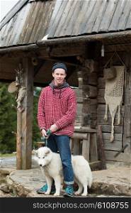 handsome young hipster portrait, young man standing together with white husky dog in front of old vintage retro wooden house