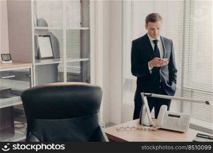 Handsome young male manager in corporate clothing, browses web pages and chats online with smart phone, has concentrated look, poses in modern office interior, reads news and browses information