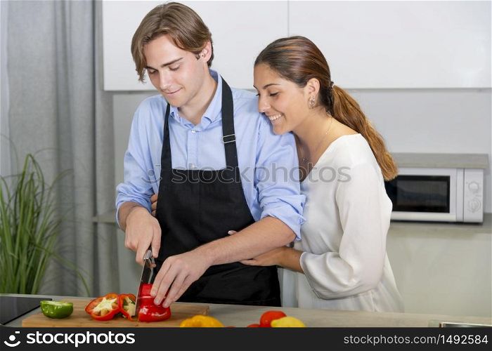 Handsome young man cutting a bell pepper while a beautiful young woman hugs him on an out of focus background. Safety and cooking at home concept.. Handsome young man cutting a bell pepper while a beautiful young woman hugs him
