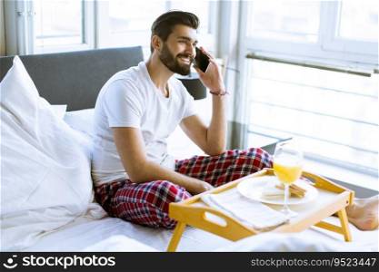 Handsome young man having breakfast in bed and using a mobile phone
