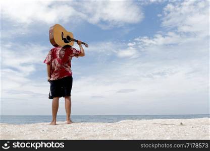 handsome young man holding guitar over shoulder on the beach with the blue sky