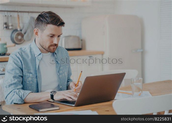 Handsome young man in casual clothes using laptop computer at home while sitting at kitchen table and remotely working from home. Freelance or distance education concept. Handsome young man using laptop computer at home