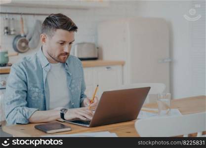 Handsome young man in casual clothes using laptop computer at home while sitting at kitchen table and remotely working from home. Freelance or distance education concept. Handsome young man using laptop computer at home