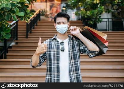 Handsome young man in protection mask holds multiple paper bag and shows his hand sign thump up. On the stairs of a shopping mall, New Normal lifestyle and shopping