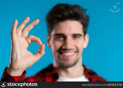 Handsome young man in red plaid shirt smiles to camera. Hipster guy showing OK sign over blue background. Winner. Success. Body language. Handsome young man in red plaid shirt smiles to camera. Hipster guy showing OK sign over blue background. Winner. Success. Body language.