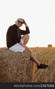 handsome young man in sunglasses, casual black sweatshirt and white short, in summer field outdoor, sitting on hay bale, haystack on sunset.. handsome young man in sunglasses, casual black sweatshirt and white short, in summer field outdoor, sitting on hay bale, haystack on sunset