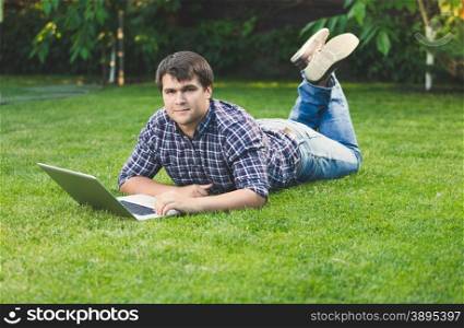 Handsome young man lying on grass at park and working with laptop