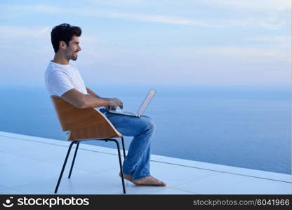 handsome young man relaxing and working on laptop computer at home balcony while looking sunset