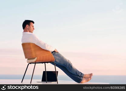 handsome young man relaxing and working on laptop computer at home balcony while looking sunset