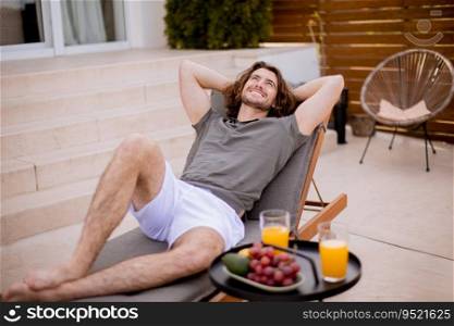 Handsome young man relaxing on deck chair with fresh fruits and cold orange juice by the swimming pool in the house backyard