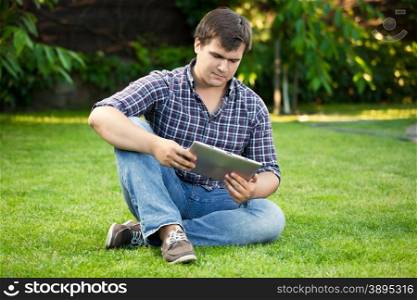 Handsome young man sitting on grass at park and using digital tablet