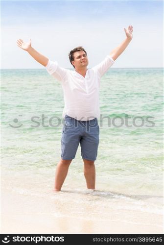 Handsome young man standing with raised hands on beach and looking in the sky
