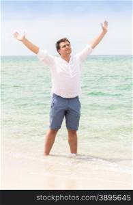 Handsome young man standing with raised hands on beach and looking in the sky