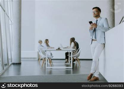 Handsome young man using his mobile phone in the office while his colleagues working in the background