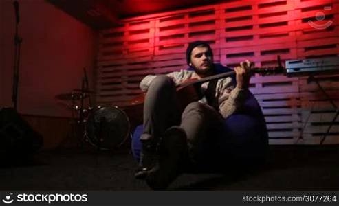 Handsome young man with beard and in hat playing acoustic guitar over musical instruments background. Stylish guitarist sitting on bean bag chair and relaxing after rehearsal with the band and picking chords on classic guitar in colorful spotlights.