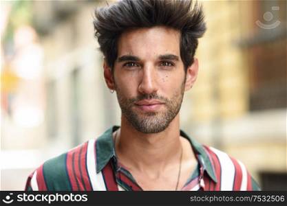 Handsome young man with dark hair and modern hairstyle wearing casual clothes in urban background.. Attractive young man with dark hair and modern hairstyle wearing casual clothes outdoors