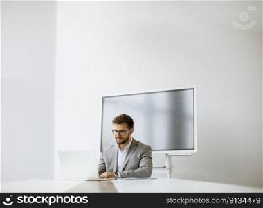 Handsome young man working on laptop in bright office with big screen behind him
