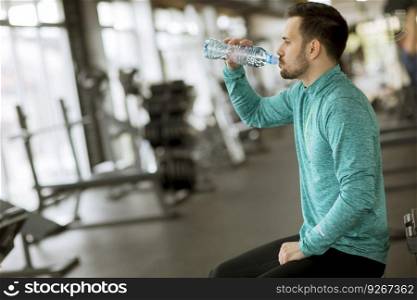 Handsome young sportsman drinking water from  bottle in the gym and resting