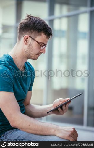 Handsome young student is sitting outdoors on the urban campus, working with a tablet pc