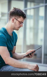 Handsome young student is sitting outdoors on the urban campus, working with a tablet pc