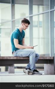 Handsome young student is sitting outdoors on the urban campus, working with a tablet pc