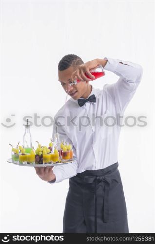 Handsome young waiter holding a tray with bottles and glass shots with grapes and mixing liquors in them on a light background. Service and vegan cocktails concept.. Young waiter holding a tray and mixing liquors