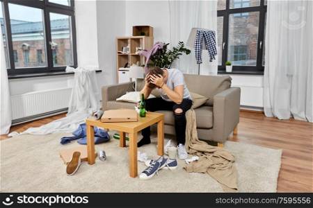 hangover, alcohol addiction and people concept - drunk man sitting on sofa in messy room at home. drunk man sitting on sofa in messy room at home