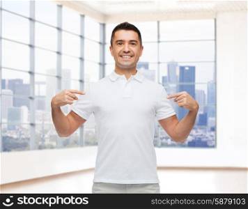 happiness, advertisement, fashion, gesture and people concept - smiling man in t-shirt pointing fingers on himself over empty apartment or office room with big window and city view background