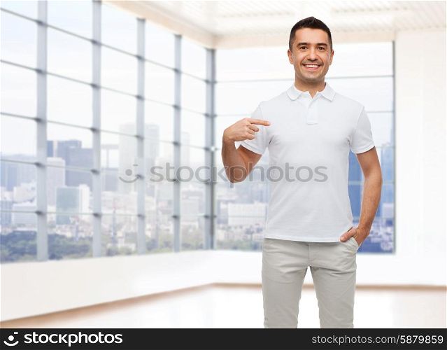 happiness, advertisement, fashion, gesture and people concept - smiling man in t-shirt pointing finger on himself over empty apartment or office room with big window and city view background