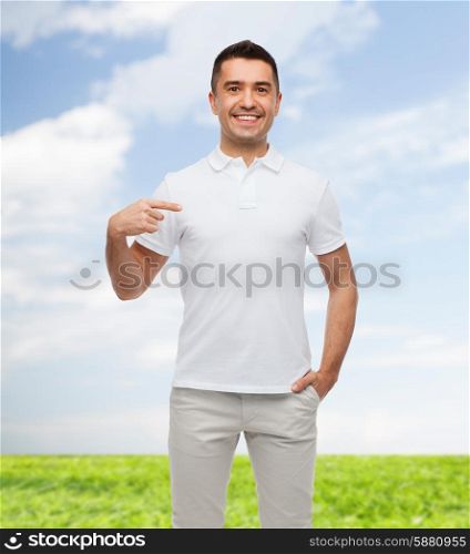 happiness, advertisement, fashion, gesture and people concept - smiling man in t-shirt pointing finger on himself over blue sky and grass background