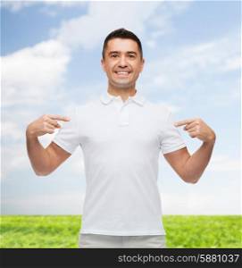 happiness, advertisement, fashion, gesture and people concept - smiling man in t-shirt pointing fingers on himself over blue sky and grass background