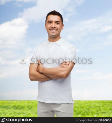 happiness and people concept - smiling man in white t-shirt with crossed arms over blue sky and grass background. smiling man in white t-shirt with crossed arms