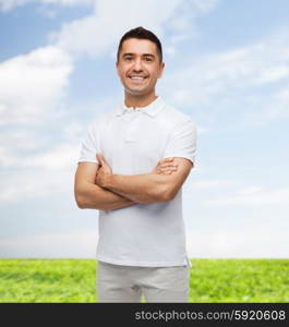 happiness and people concept - smiling man in white t-shirt with crossed arms over blue sky and grass background. smiling man in white t-shirt with crossed arms