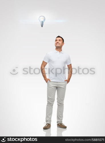 happiness and people concept - smiling man with hands in pockets looking up to lighting bulb over gray background