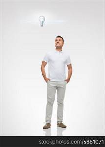 happiness and people concept - smiling man with hands in pockets looking up to lighting bulb over gray background