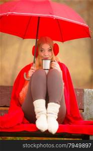 Happiness carefree and fall concept. Young woman in red relaxing in autumn park on bench under umbrella, enjoying hot drink holding mug with warm beverage. Orange leaves background