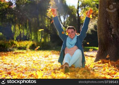 Happiness carefree. Young pretty woman relaxing in the autumn park throwing leaves up in the air with arms raised up, smiling elated expression. Beautiful girl in colorful forest foliage outdoor.