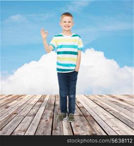 happiness, childhood and people concept - smiling little boy in casual clothes making ok gesture over blue sky and wooden floor background