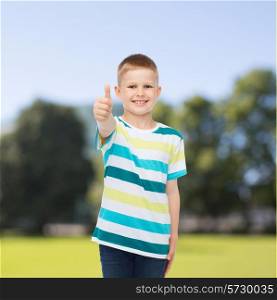 happiness, childhood, nature and people concept - smiling little boy in casual clothes showing thumbs up over green park background
