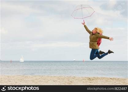 Happiness, enjoying cold autumn weather, feeling great concept. Woman jumping with transparent umbrella on beach near sea, sunny day and clear blue sky. Woman jumping with transparent umbrella on beach