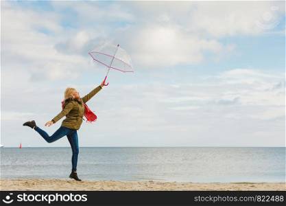 Happiness, enjoying cold autumn weather, feeling great concept. Woman jumping with transparent umbrella on beach near sea, sunny day and clear blue sky. Woman jumping with transparent umbrella on beach