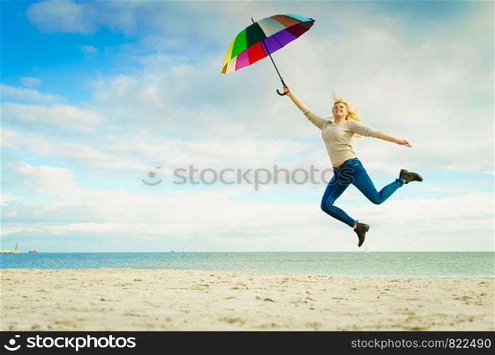 Happiness, enjoying weather, feeling great concept. Woman jumping with colorful umbrella on beach near sea, sunny day and clear blue sky. Woman jumping with colorful umbrella on beach