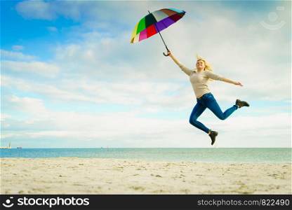 Happiness, enjoying weather, feeling great concept. Woman jumping with colorful umbrella on beach near sea, sunny day and clear blue sky. Woman jumping with colorful umbrella on beach