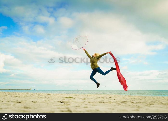 Happiness, enjoying weather, feeling great concept. Woman jumping with red scarf and transparent umbrella on beach near sea, sunny day, blue with clouds sky. Happy woman jumping with scarf on beach