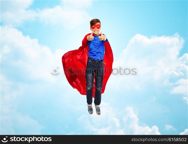 happiness, freedom, childhood, movement and people concept - boy in red super hero cape and mask flying in air and showing thumbs up over blue sky and clouds background