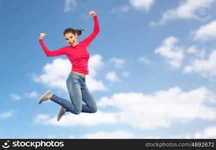 happiness, freedom, power, motion and people concept - smiling young woman jumping in air with raised fists over blue sky and clouds background