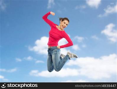 happiness, freedom, power, motion and people concept - smiling young woman jumping in air with raised fist over blue sky and clouds background. smiling young woman jumping in air