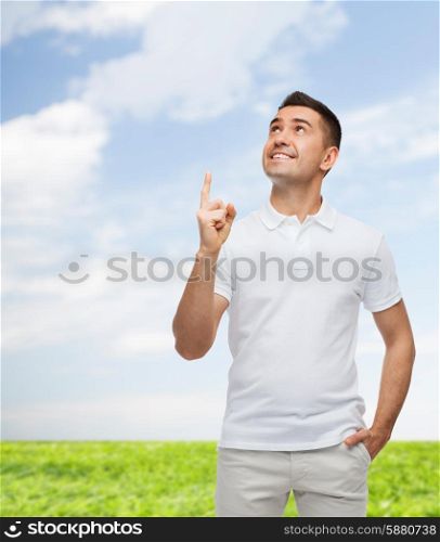 happiness, gesture and people concept - smiling man pointing finger up over blue sky and grass background