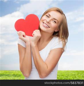 happiness, health, people, holidays and love concept - smiling young woman in white t-shirt holding red heart over blue sky and grass background